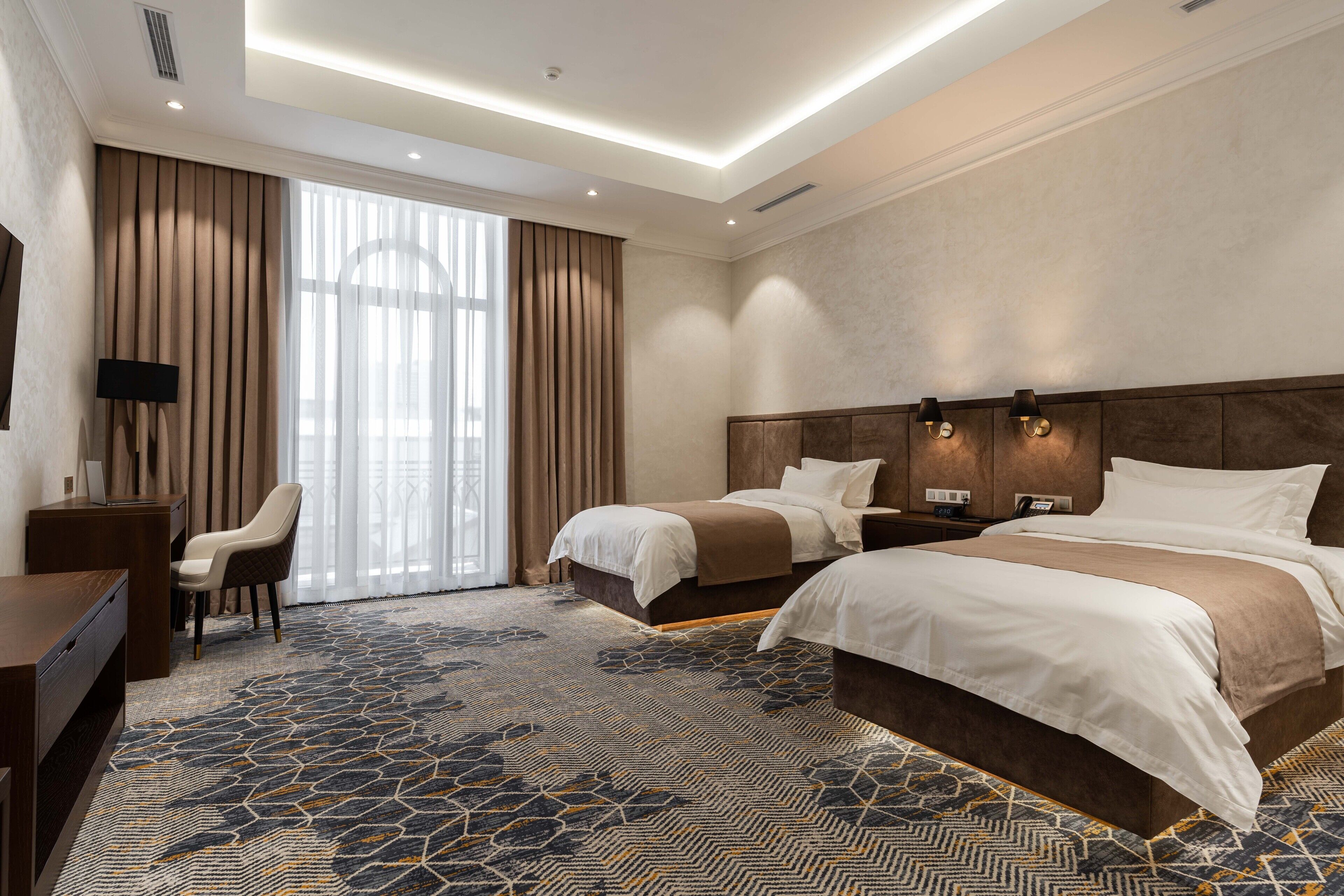 A hotel room with two beds, a desk, a chair, and a large window with curtains.