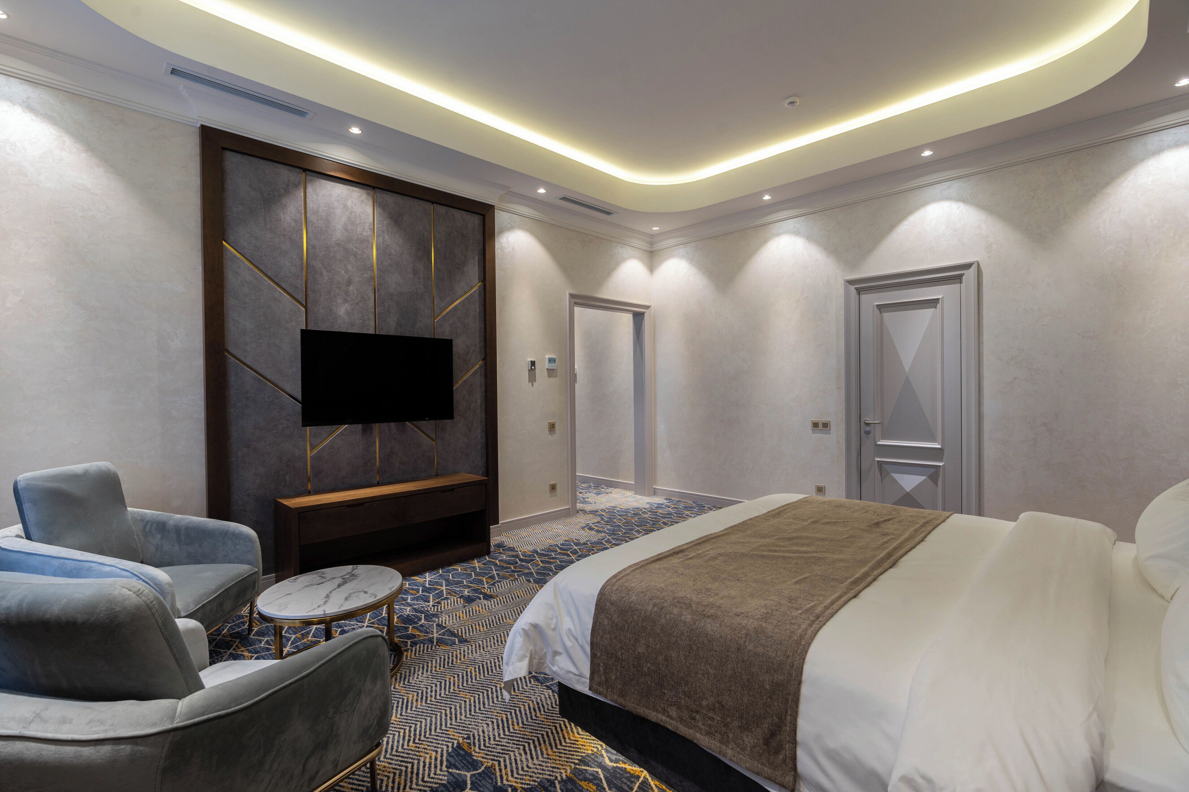 A modern hotel room with a large bed, a flat-screen TV mounted on a wall, a seating area with a sofa and coffee table, and a patterned carpet.
