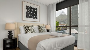 A modern hotel room with a large bed, two bedside tables with lamps, a framed abstract artwork, and a view of a mountain through a window.