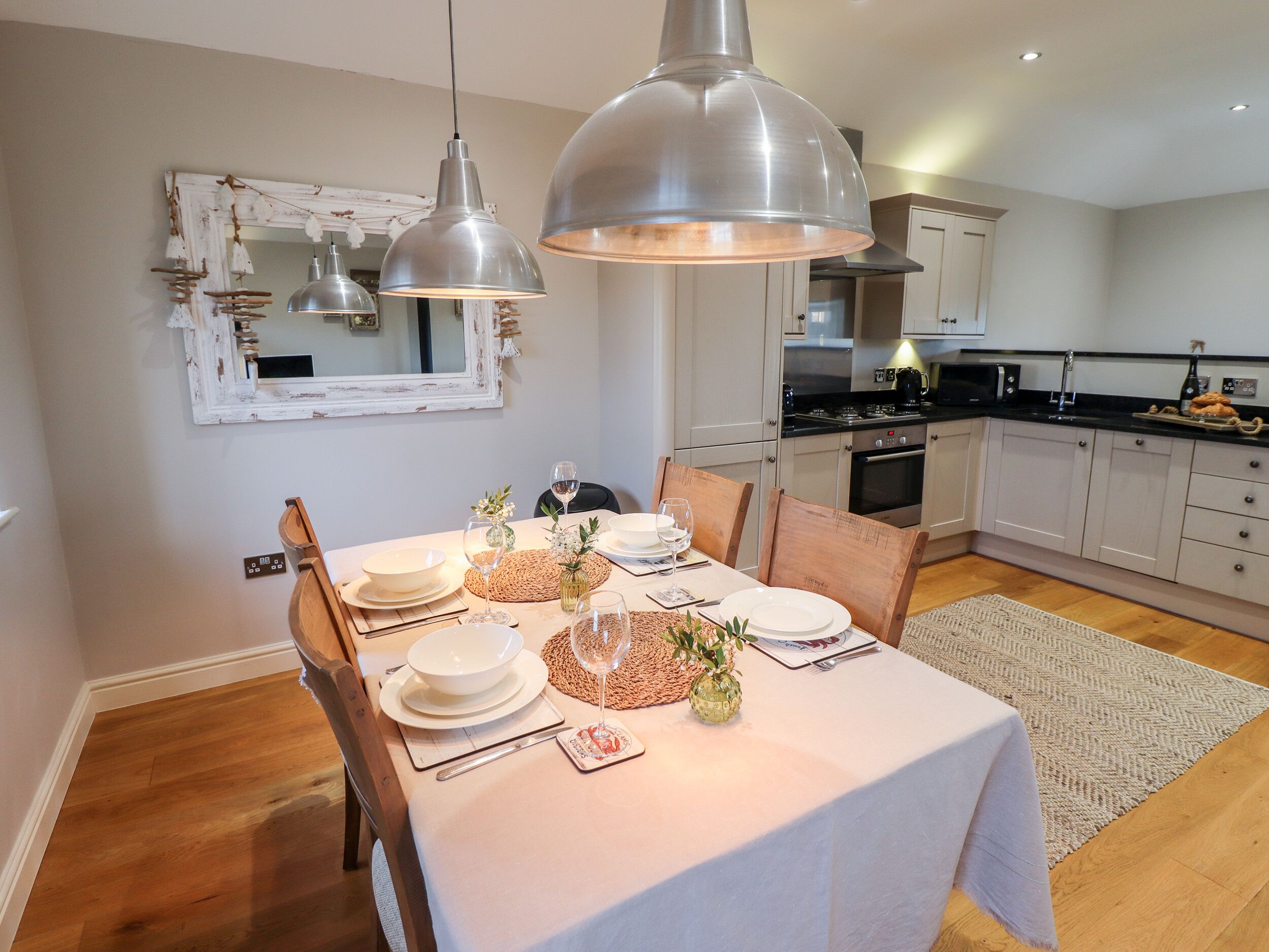 Uma sala de jantar com uma mesa posta para quatro pessoas, uma cozinha ao fundo e luminárias pendentes sobre a mesa.