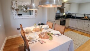 Comedor con una mesa dispuesta para cuatro, cocina al fondo e iluminación colgante sobre la mesa.