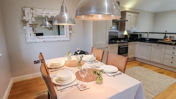 Uma sala de jantar com uma mesa posta para quatro pessoas, uma cozinha ao fundo e luminárias pendentes sobre a mesa.