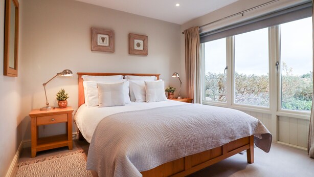 Un dormitorio con una cama grande, dos mesitas de noche, un ventanal con cortinas y dos cuadros enmarcados en la pared.