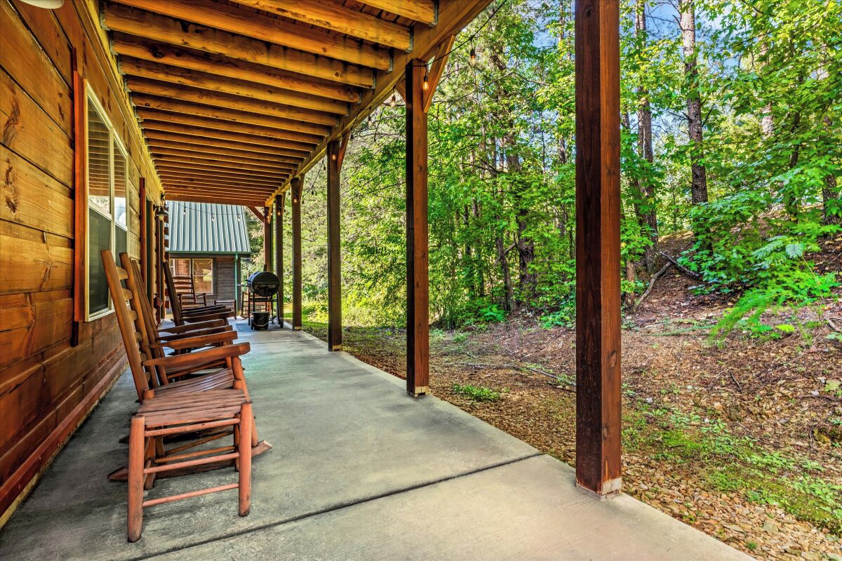 A covered porch with wooden chairs and a concrete floor, surrounded by a forested area.