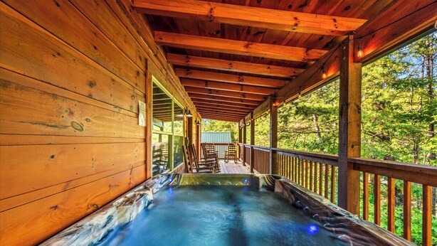 A wooden cabin with a hot tub under a covered porch, surrounded by trees.
