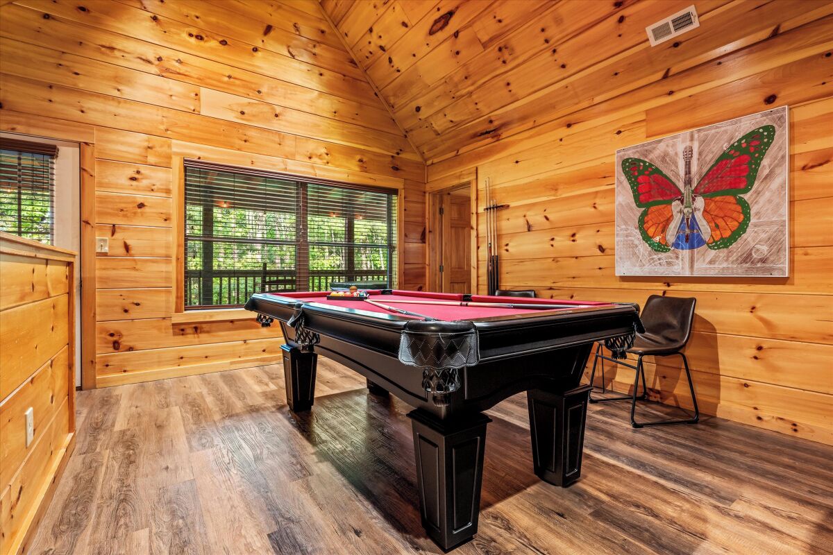 A wooden room with a pool table, a window with blinds, and a butterfly painting on the wall.