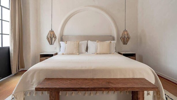 Une chambre avec un grand lit, un banc en bois, deux tables de chevet et une grande fenêtre en arc de cercle.