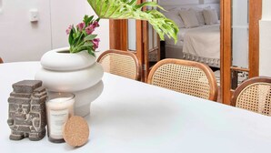 Un coin repas avec une table blanche, des chaises en osier, un vase de fleurs, une bougie et une pierre décorative.