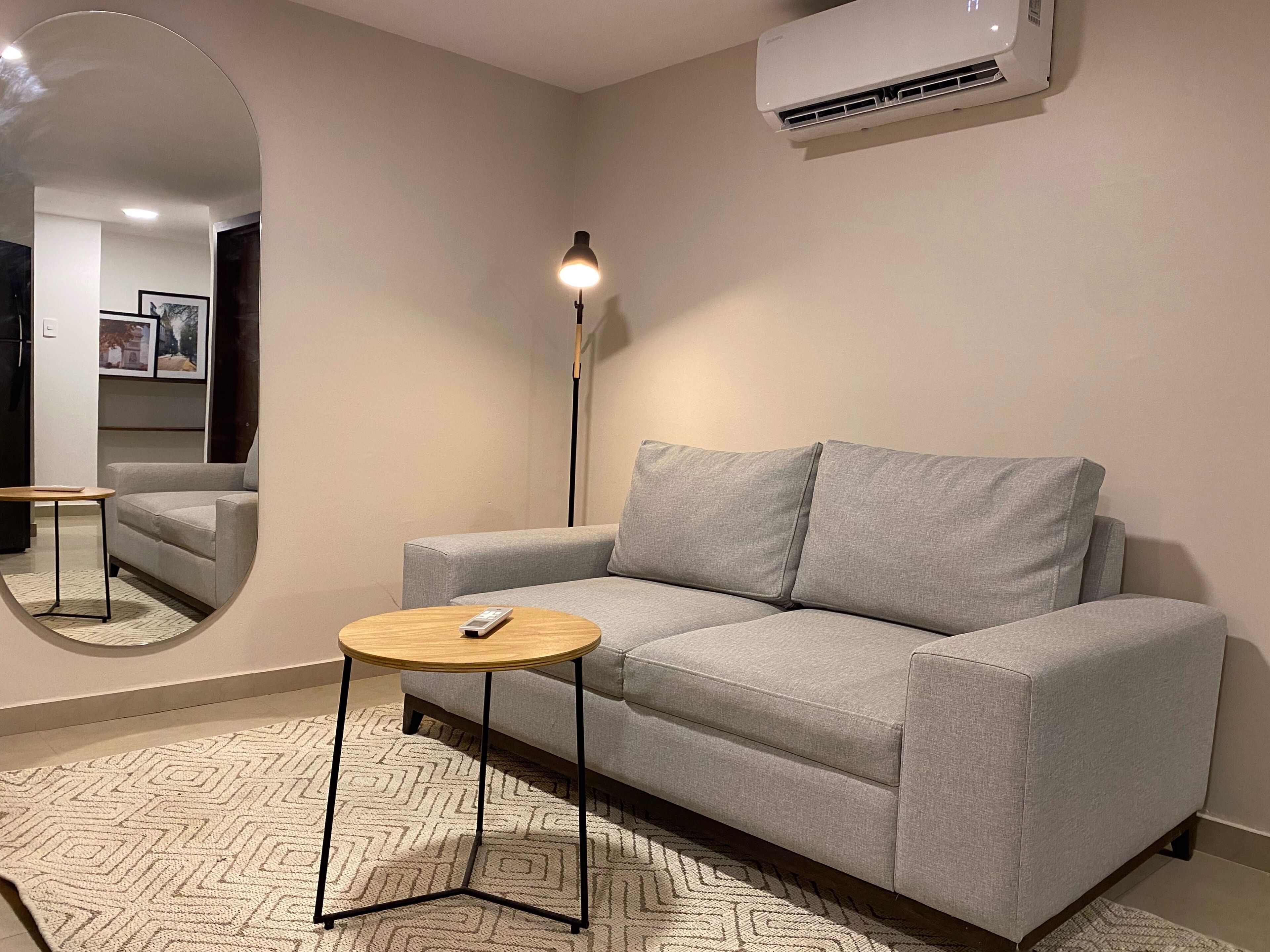 A modern living room with a grey sofa, a small round table, a floor lamp, and a large mirror.
