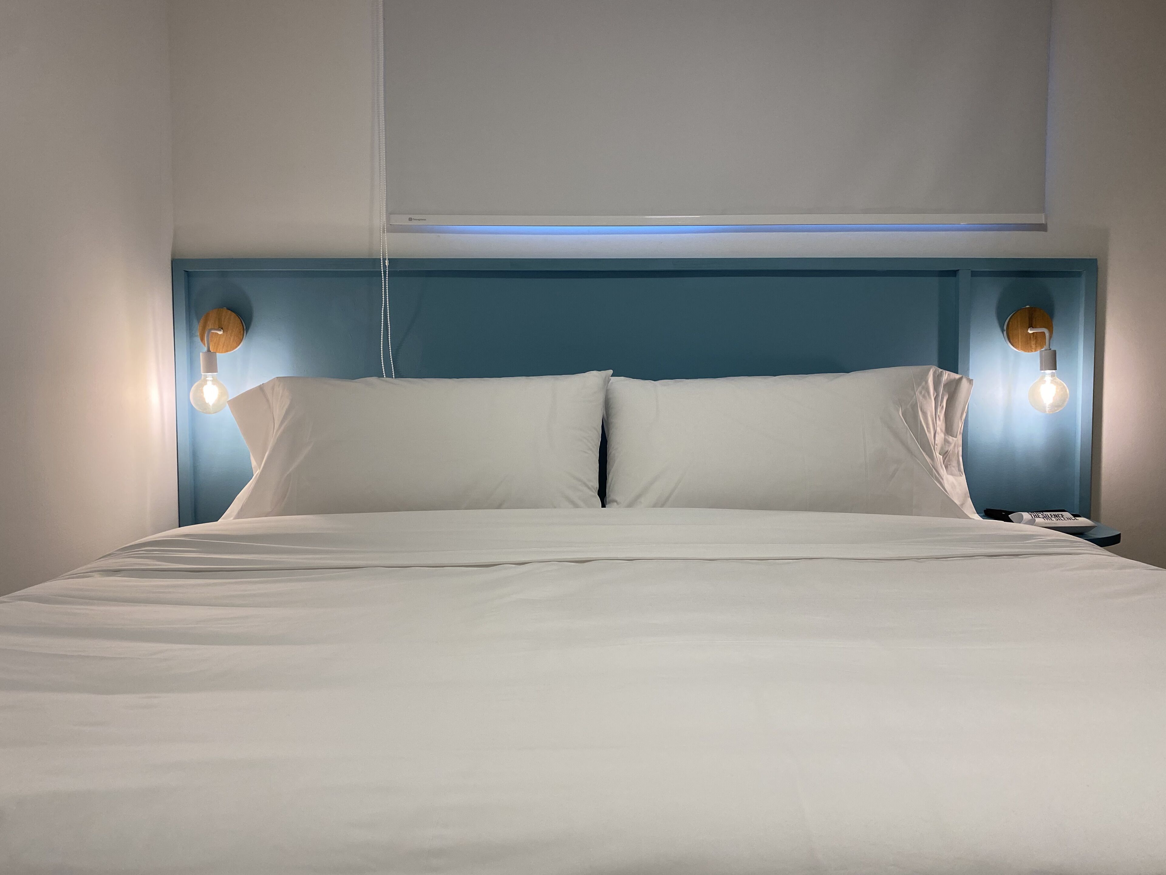A neatly made bed with white linens, a blue headboard, and two wall-mounted light fixtures.