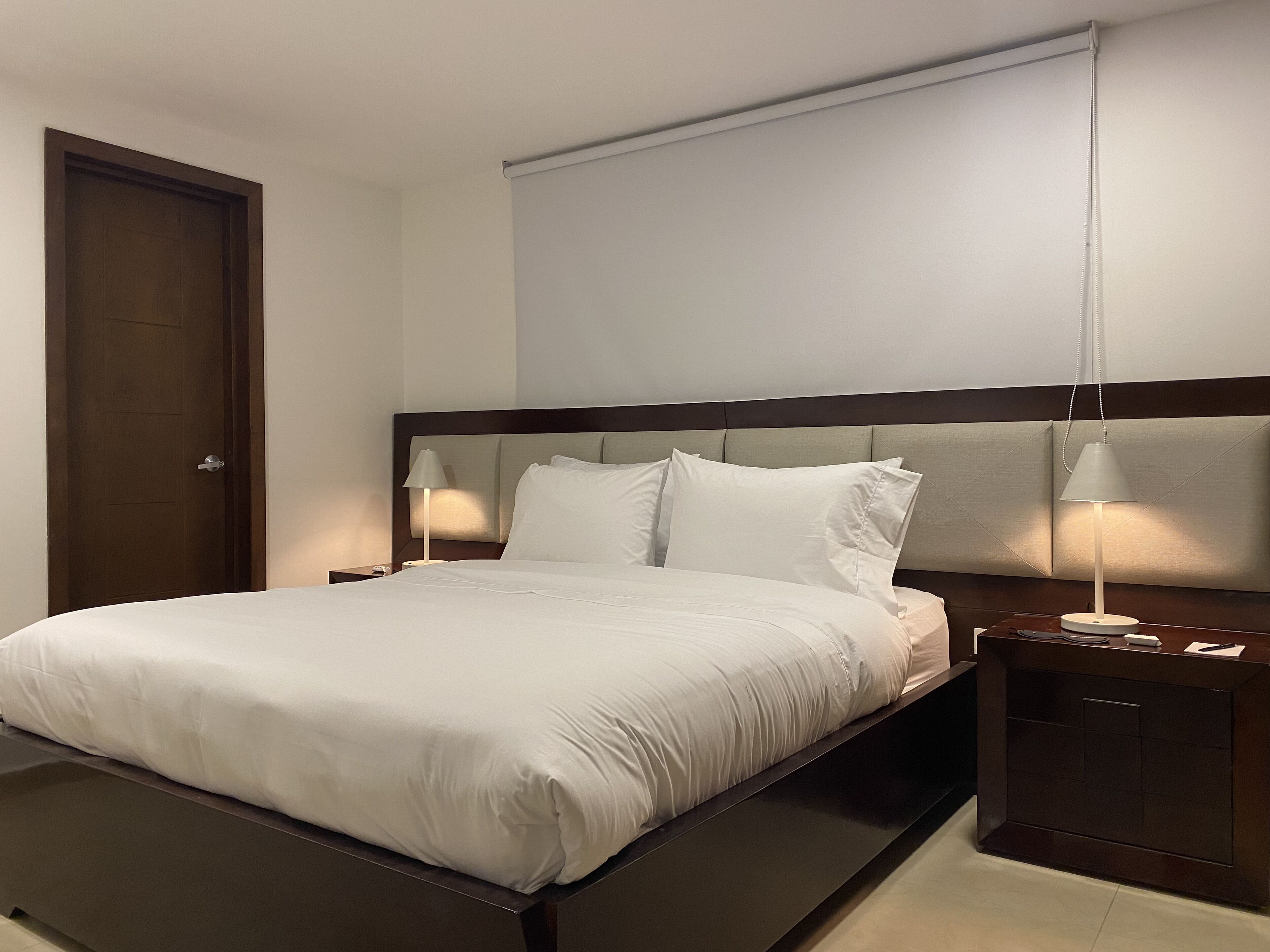 A neatly made bed with white linens, a wooden headboard, two bedside tables with lamps, and a closed door.