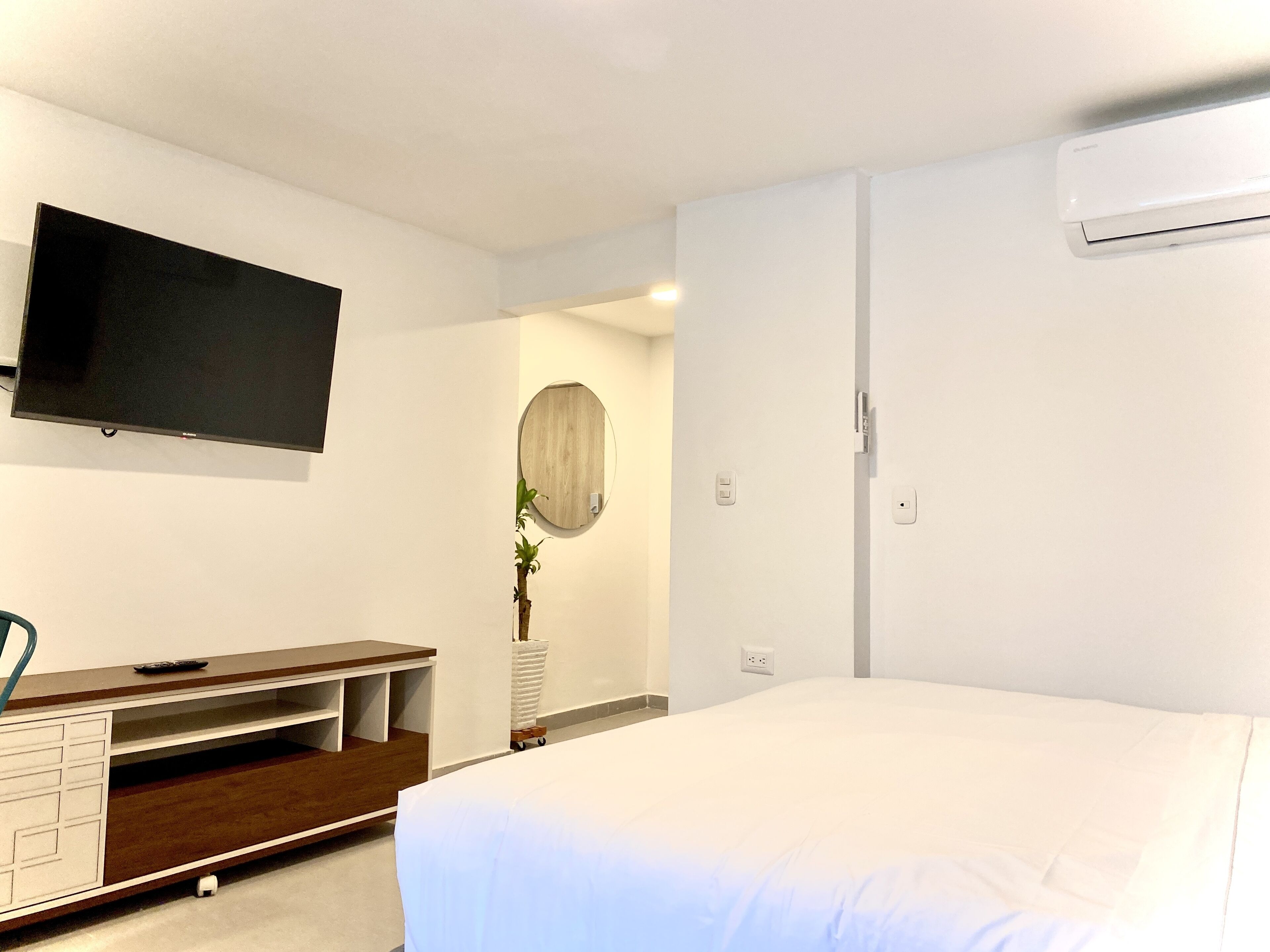 A bedroom with a bed, a TV mounted on the wall, a wooden dresser, a mirror, and a wall-mounted air conditioner.