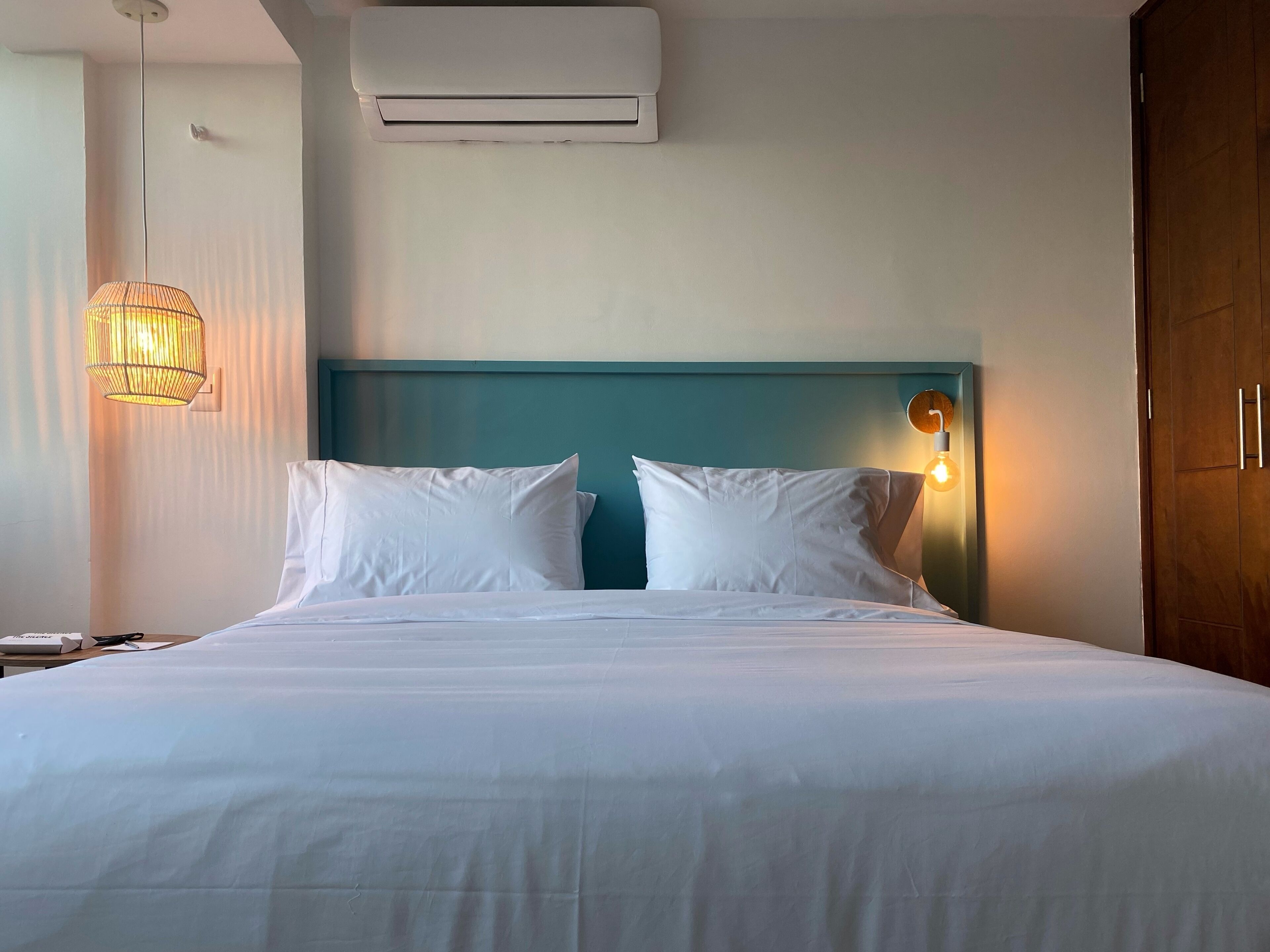 A neatly made bed with white linens, a teal headboard, and a wall-mounted air conditioner above it.