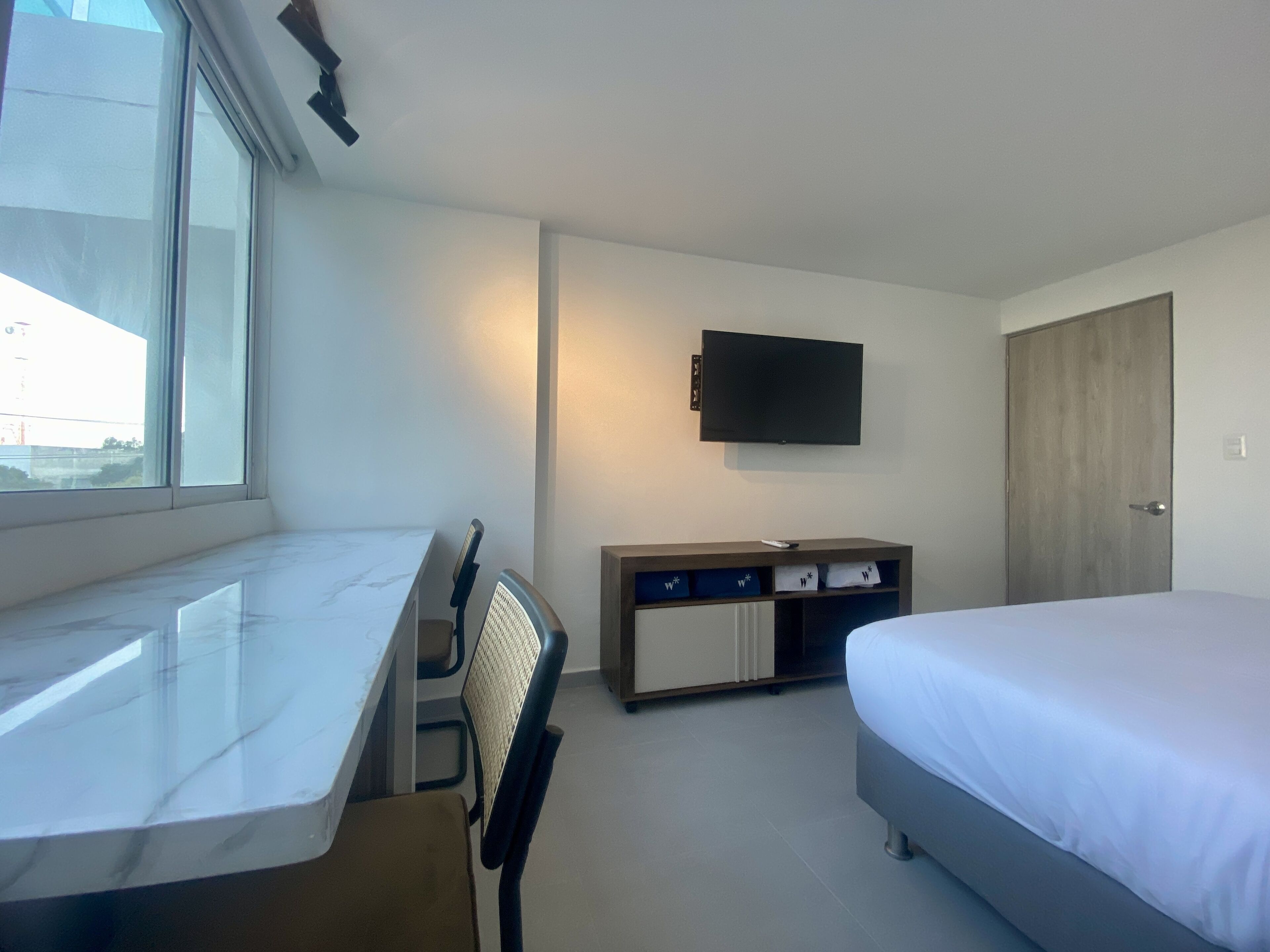 A hotel room with a bed, a desk with a chair, a TV mounted on the wall, and a wardrobe.