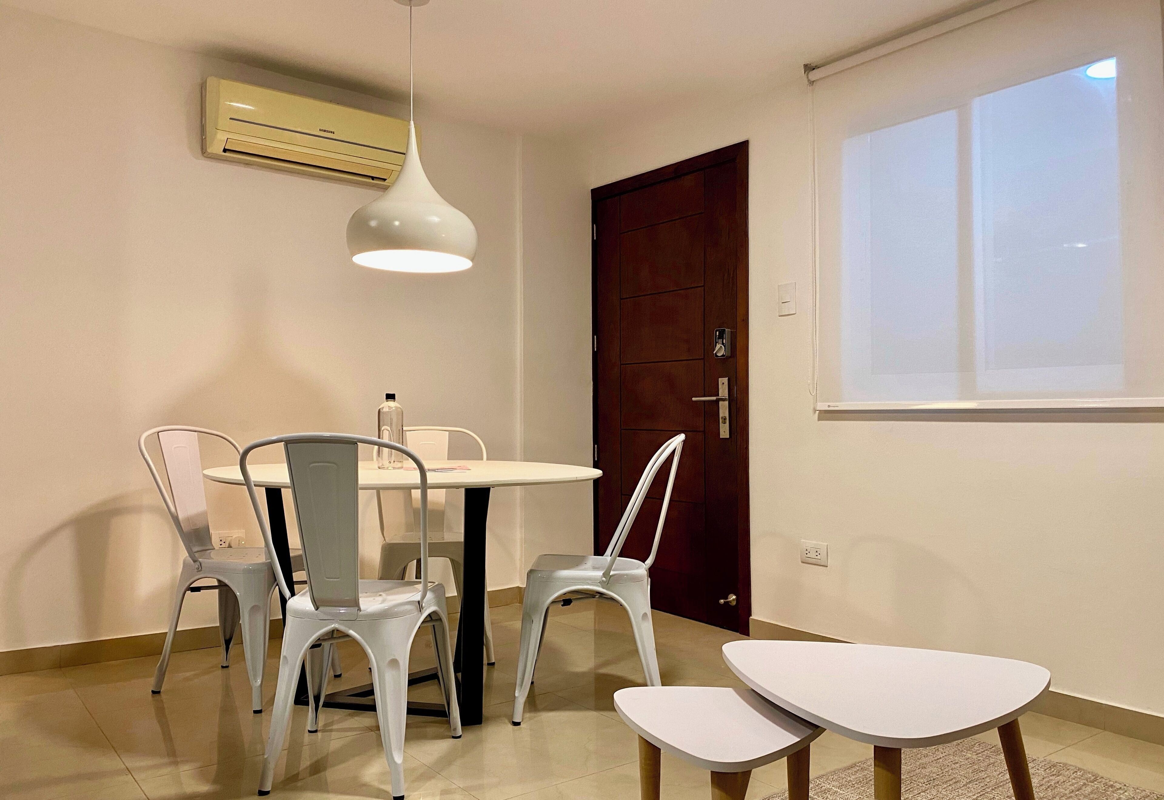 A dining area with a round table and four chairs, an air conditioner, a white pendant light, and a window with blinds.