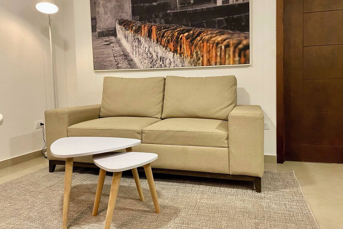 Un salon avec un canapé beige, deux petites tables d’appoint blanches, un lampadaire et un tableau encadré représentant un tigre accroché au mur.