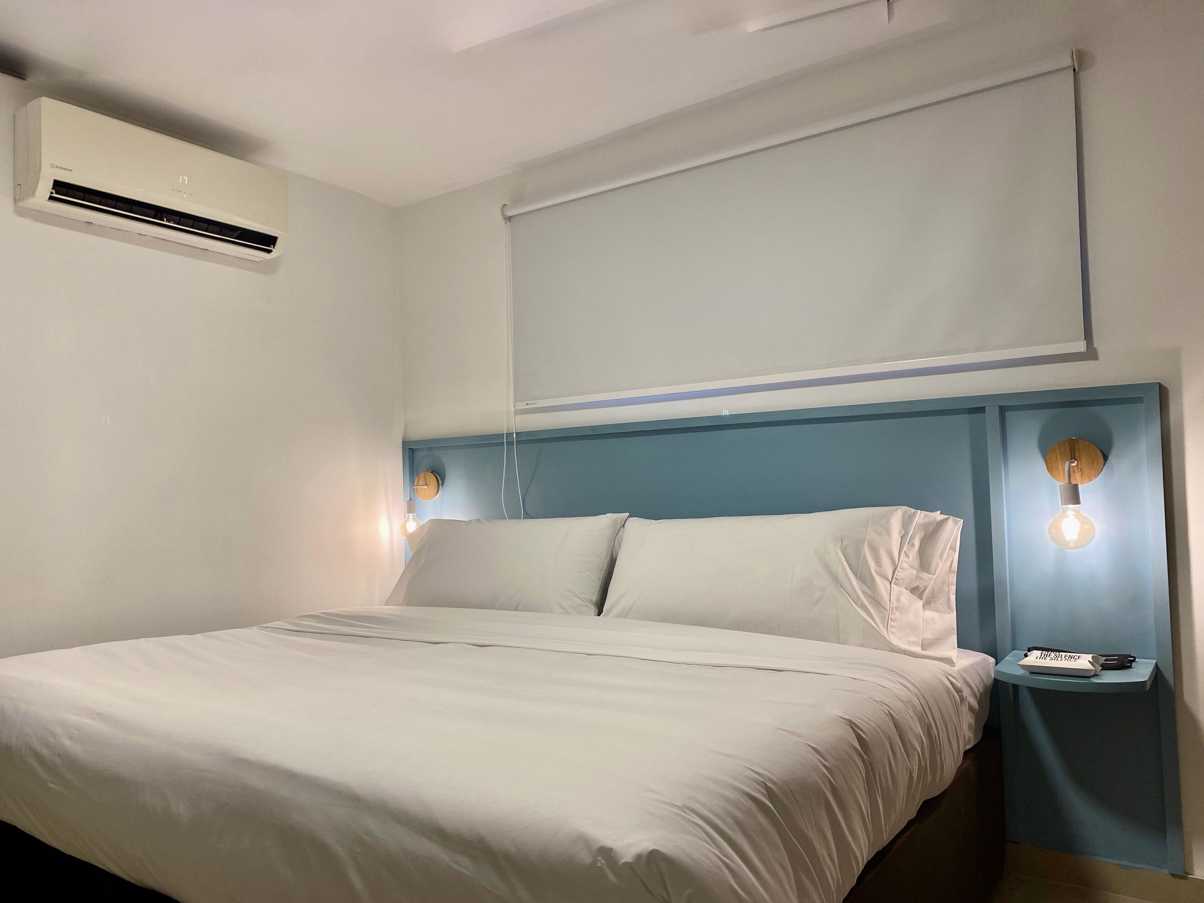 Habitación de hotel con cama, mesita de noche, aire acondicionado instalado en la pared y ventana con persiana.