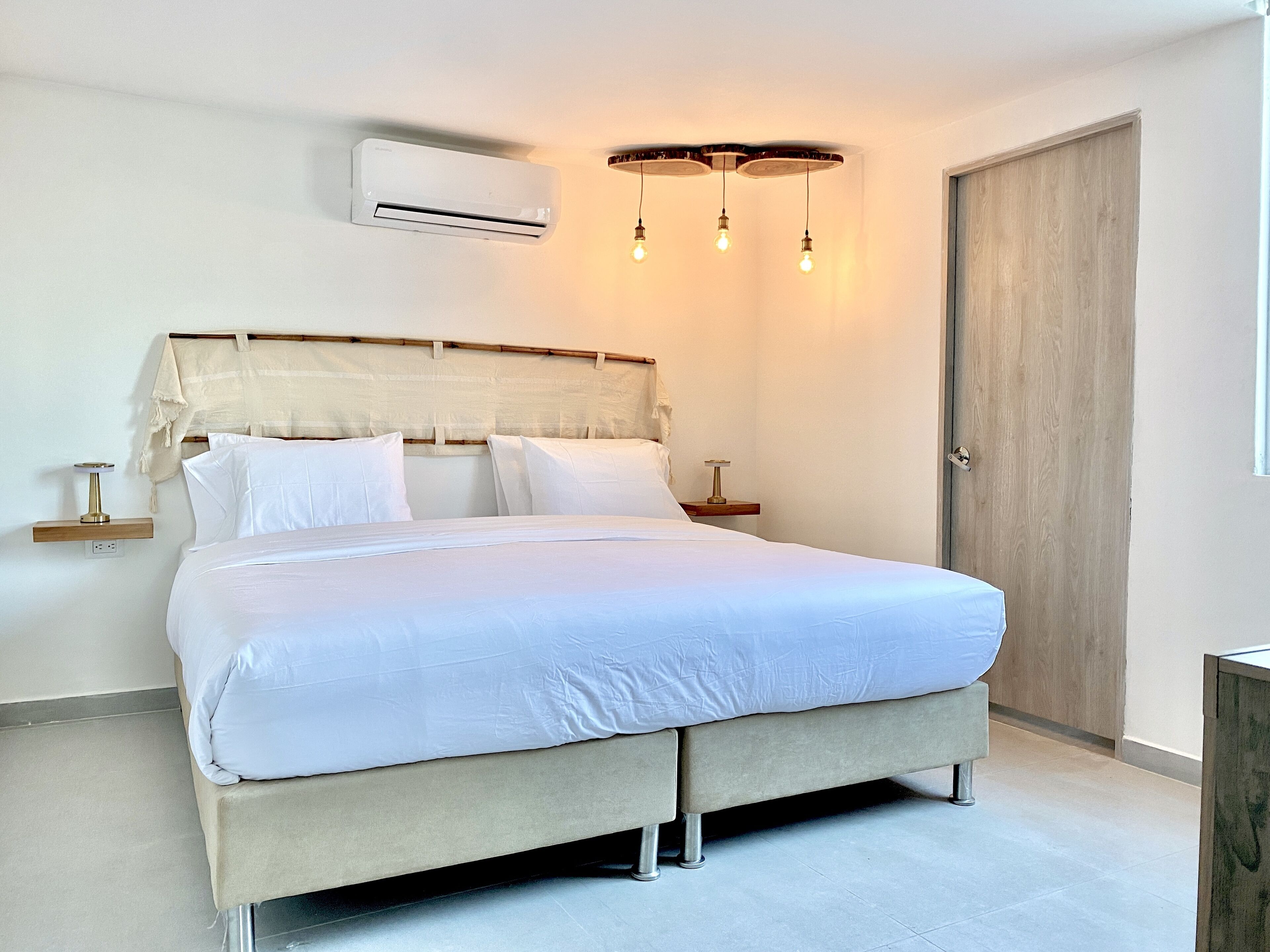 A neatly made bed with a headboard, an air conditioning unit above it, and a bedside table with a lamp.