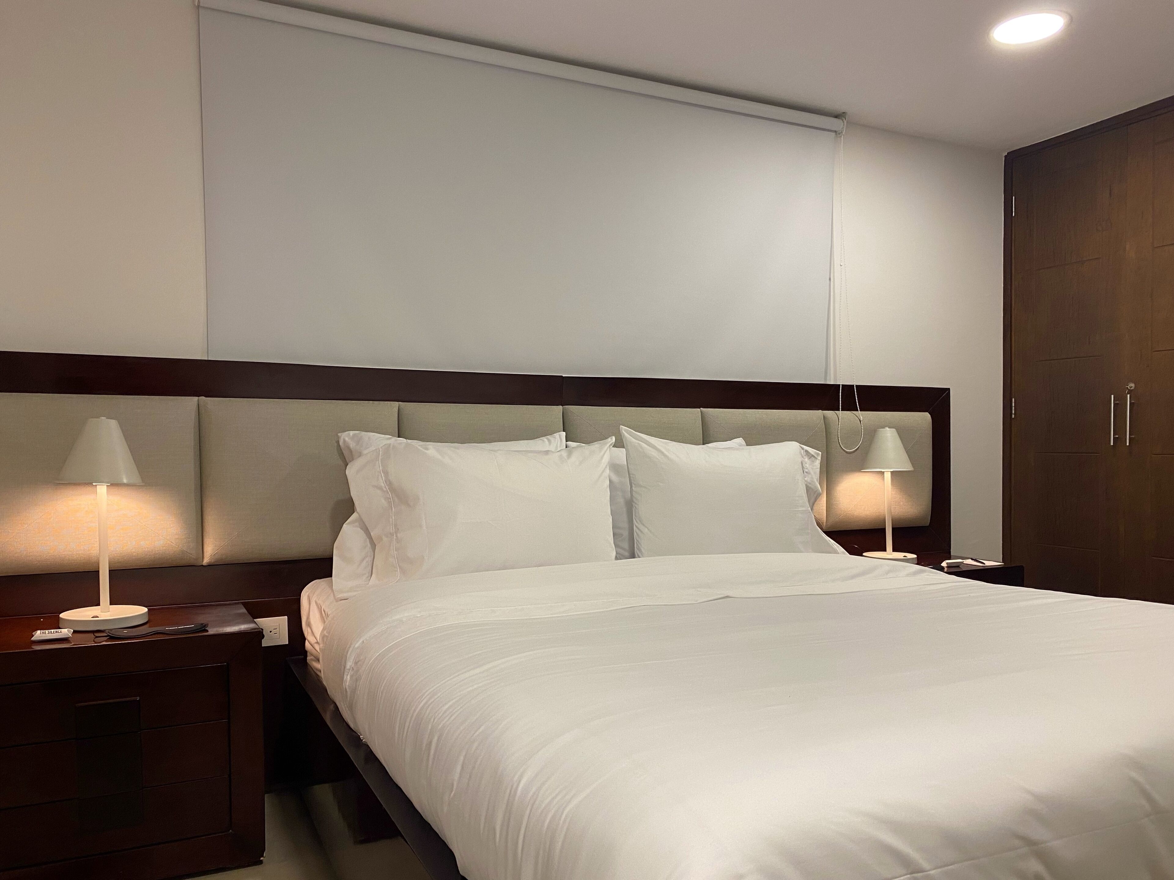 A neatly made bed with white linens, two bedside lamps, a wooden headboard, and a wooden wardrobe.