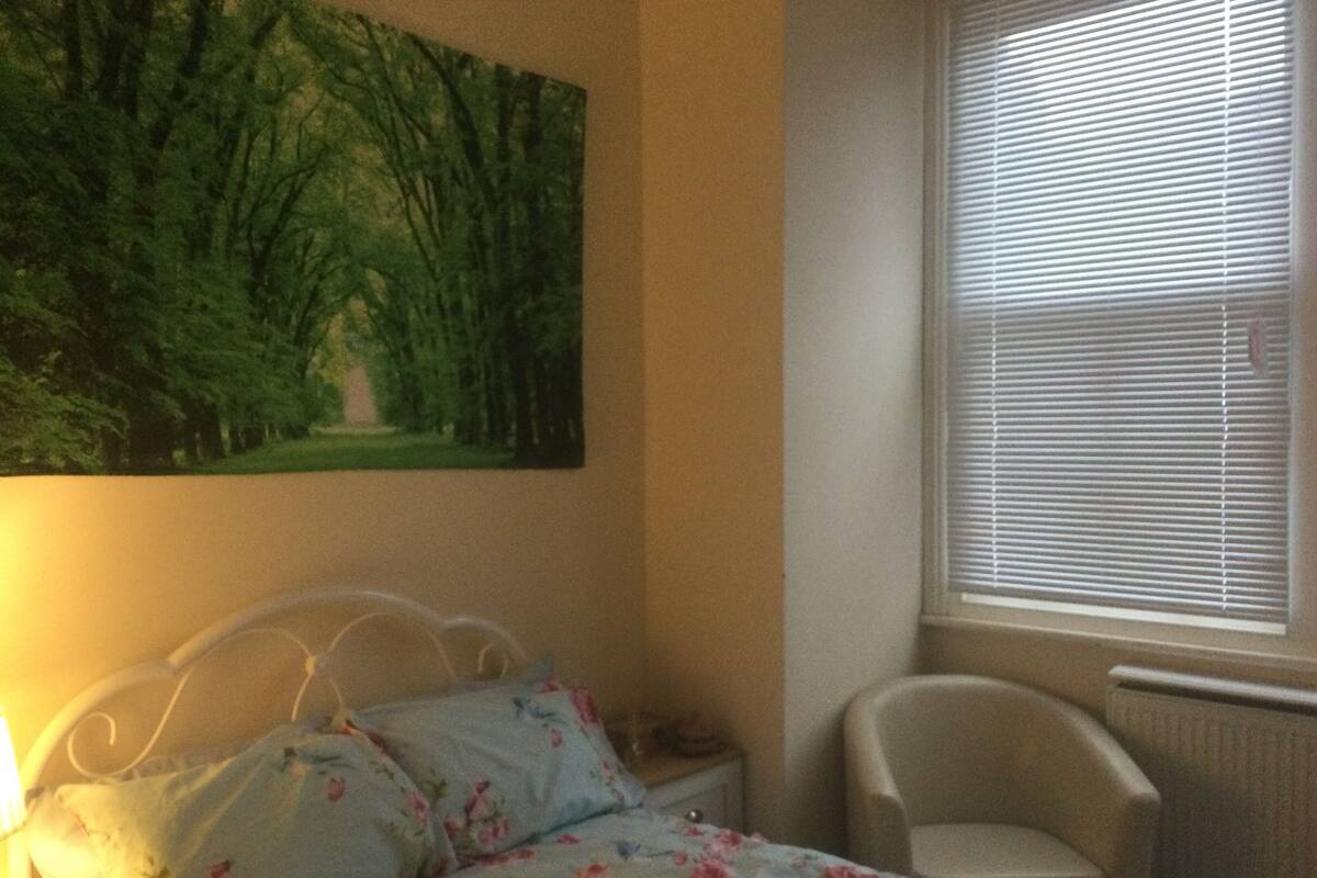 A bedroom with a bed, a chair, a window with blinds, and a painting on the wall.