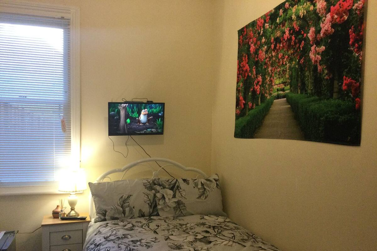 A bedroom with a bed, a TV on the wall, a window with blinds, and a painting of a garden.