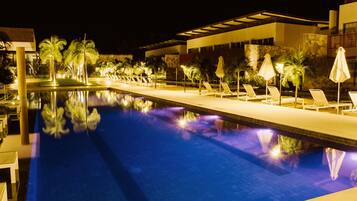 A nighttime swimming pool with lounge chairs and umbrellas, surrounded by palm trees and modern architecture.