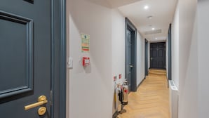 A hallway with dark wooden doors, white walls, and a wooden floor. There are fire extinguishers and a fire alarm on the wall.