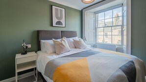 A bedroom with a large bed, a nightstand, a wall-mounted lamp, and a window with a view of buildings. - Priorsgate | Oliphant Apartment (St Andrews)