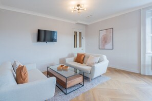 A modern living room with a sofa, armchair, glass coffee table, TV, and artwork on the wall. - Priorsgate | Oliphant Apartment (St Andrews)