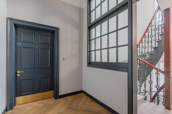 Un couloir avec une porte en bois sombre, une fenêtre à carreaux et un escalier en bois orné de rampes en métal ouvragé.