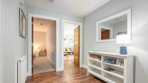 Un couloir doté d’un plancher en bois, de murs blancs et d’une console blanche sur laquelle sont posés une lampe et des livres.