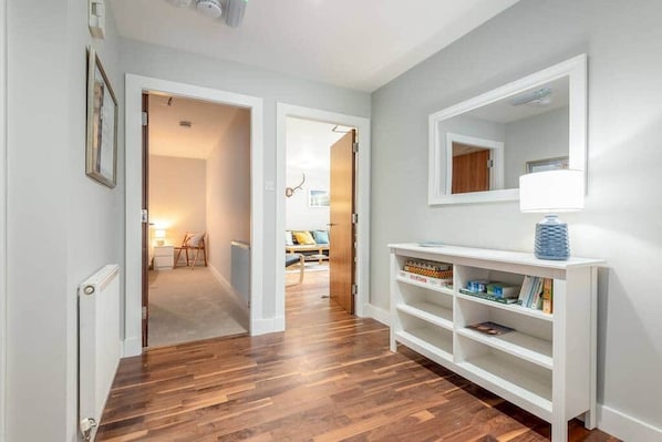 A hallway with wooden flooring, white walls, and a white console table with a lamp and books. - Southfield Apartments (No 14) (St Andrews)