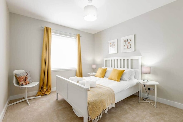 A bedroom with a white bed, yellow curtains, a bedside table with a lamp, and a small chair.