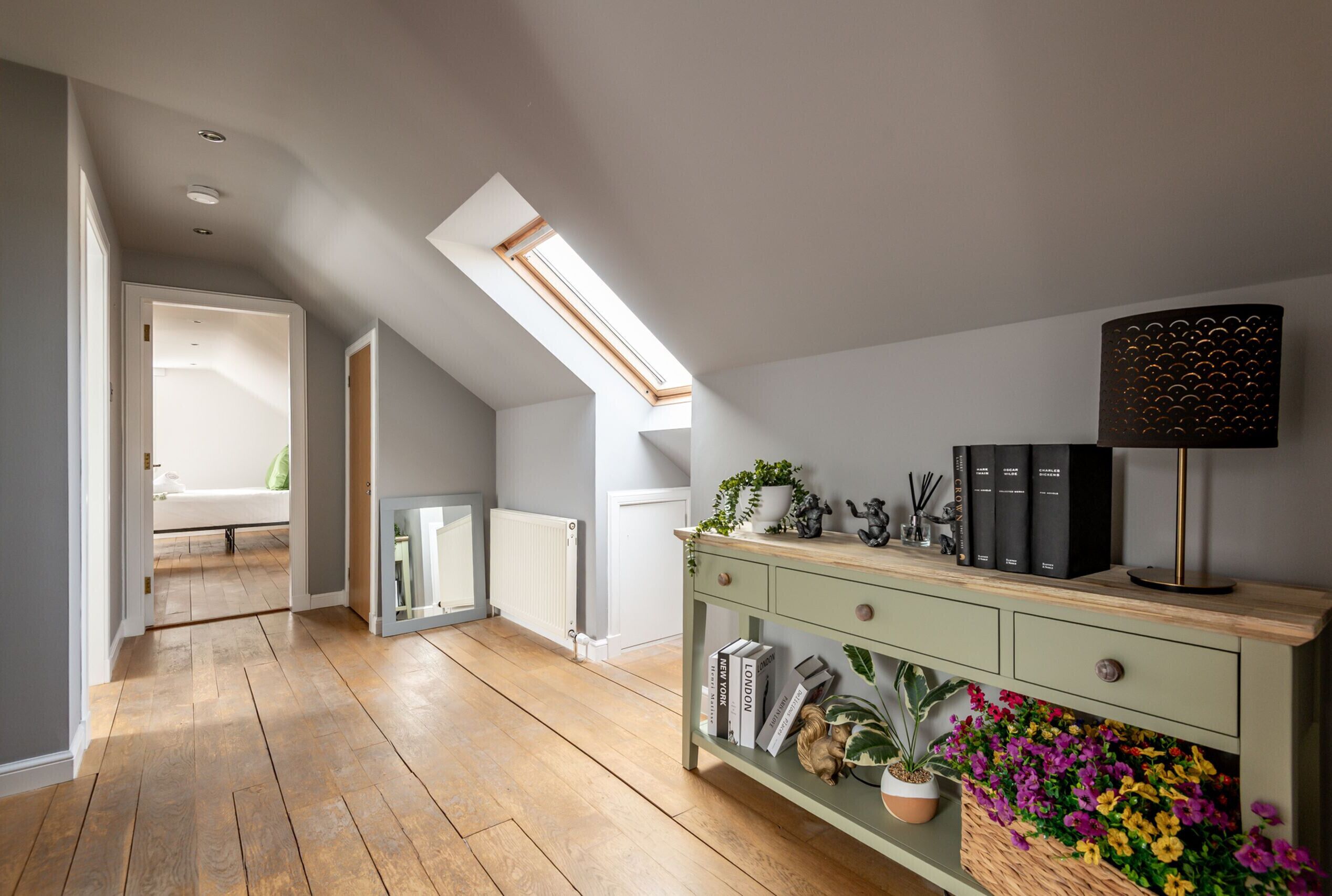 Une pièce avec un plafond en pente, un puits de lumière, un plancher en bois, une console verte ornée de livres et d’objets décoratifs, et une vue sur une pièce blanche à l’arrière-plan.