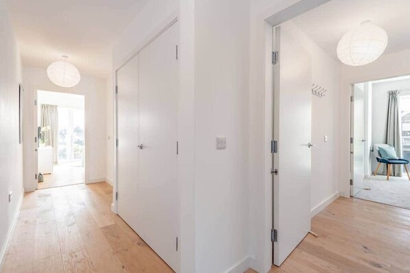 A hallway with white walls and wooden flooring, leading to a room with a large window and a chair.