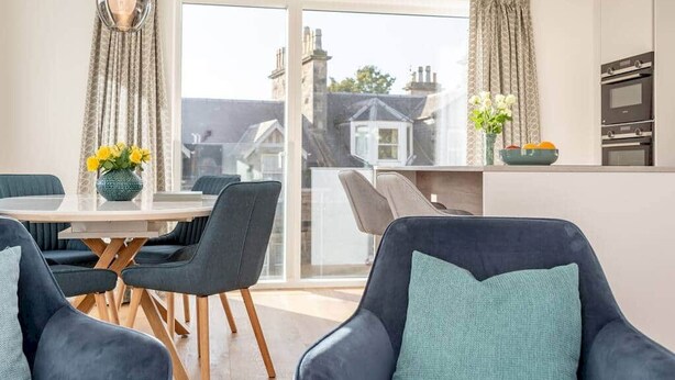 A modern living room with a round table, dark sofas, and a view of the city through large windows. - Lade Braes Apartments (No 22D) (St Andrews)