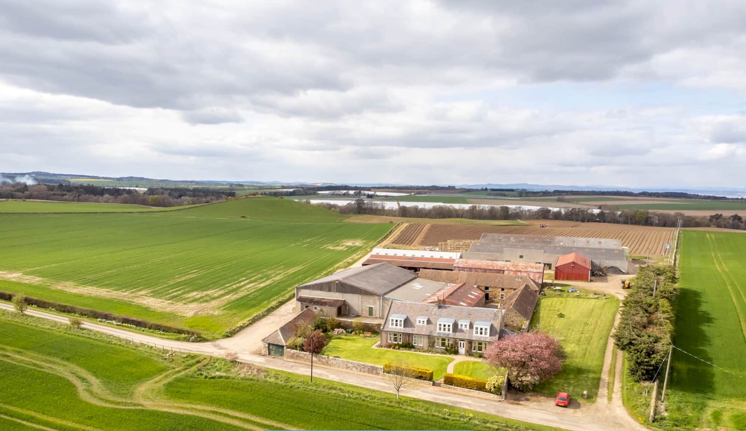 A large farmstead with multiple buildings, surrounded by expansive green fields and a clear sky.
