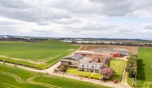 A large farmstead with multiple buildings, surrounded by expansive green fields and a clear sky.