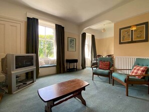 Ein Wohnzimmer mit Fernseher, einer Holzbank, einem türkisfarbenen Sessel und einem Fenster mit Vorhängen.