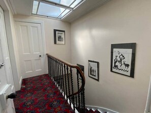 Ein Flur mit einem gemusterten Teppich, einer weißen Tür, einer Treppe mit einem Holzgeländer und gerahmten Kunstwerken an den Wänden.