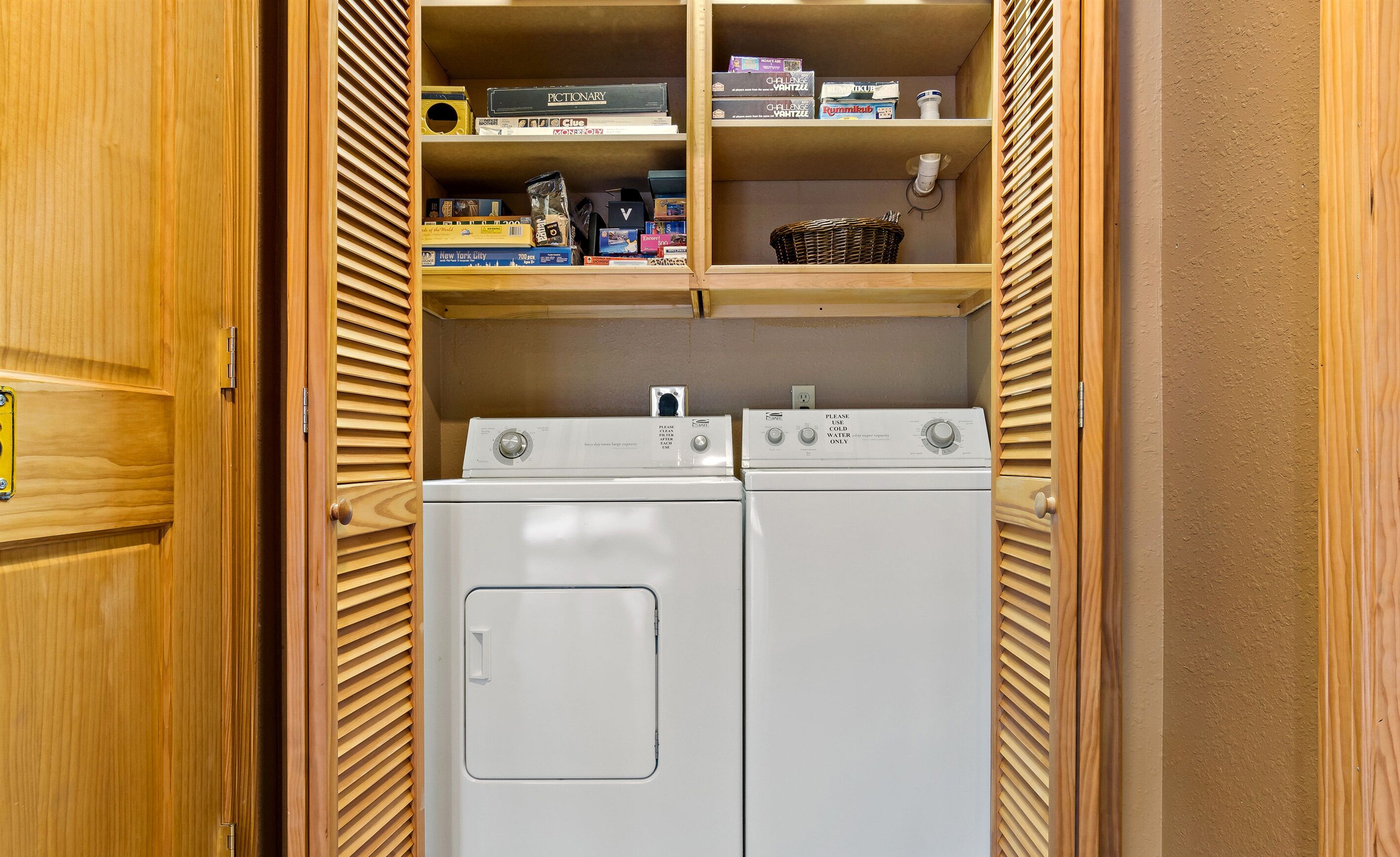 A laundry room with two front-loading washing machines and shelves above containing various items.