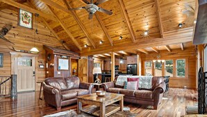 Ein geräumiges Blockhaus-Interieur mit einem Wohnzimmer, das zwei Ledersofas, einen Holztisch und einen Deckenventilator zeigt.