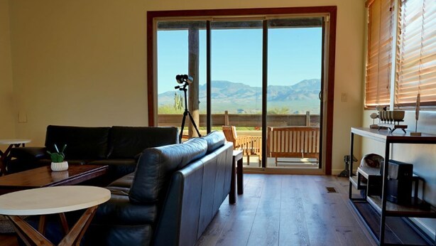 A living room with a black leather sofa, a wooden coffee table, and a sliding glass door leading to a balcony with a tripod and a view of mountains.