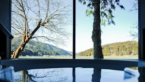Ein Zimmer mit großem Fenster und Blick auf einen See, Bäume und einen bewaldeten Hügel.
