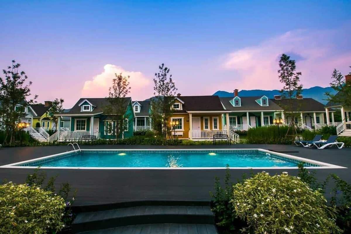 A row of houses with a central swimming pool, surrounded by greenery and a mountainous backdrop.