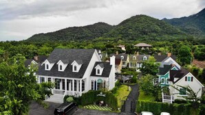 A residential area with houses featuring gabled roofs and white exteriors, surrounded by lush greenery and hills in the background. - Vermont Khao Yai (Pak Chong)