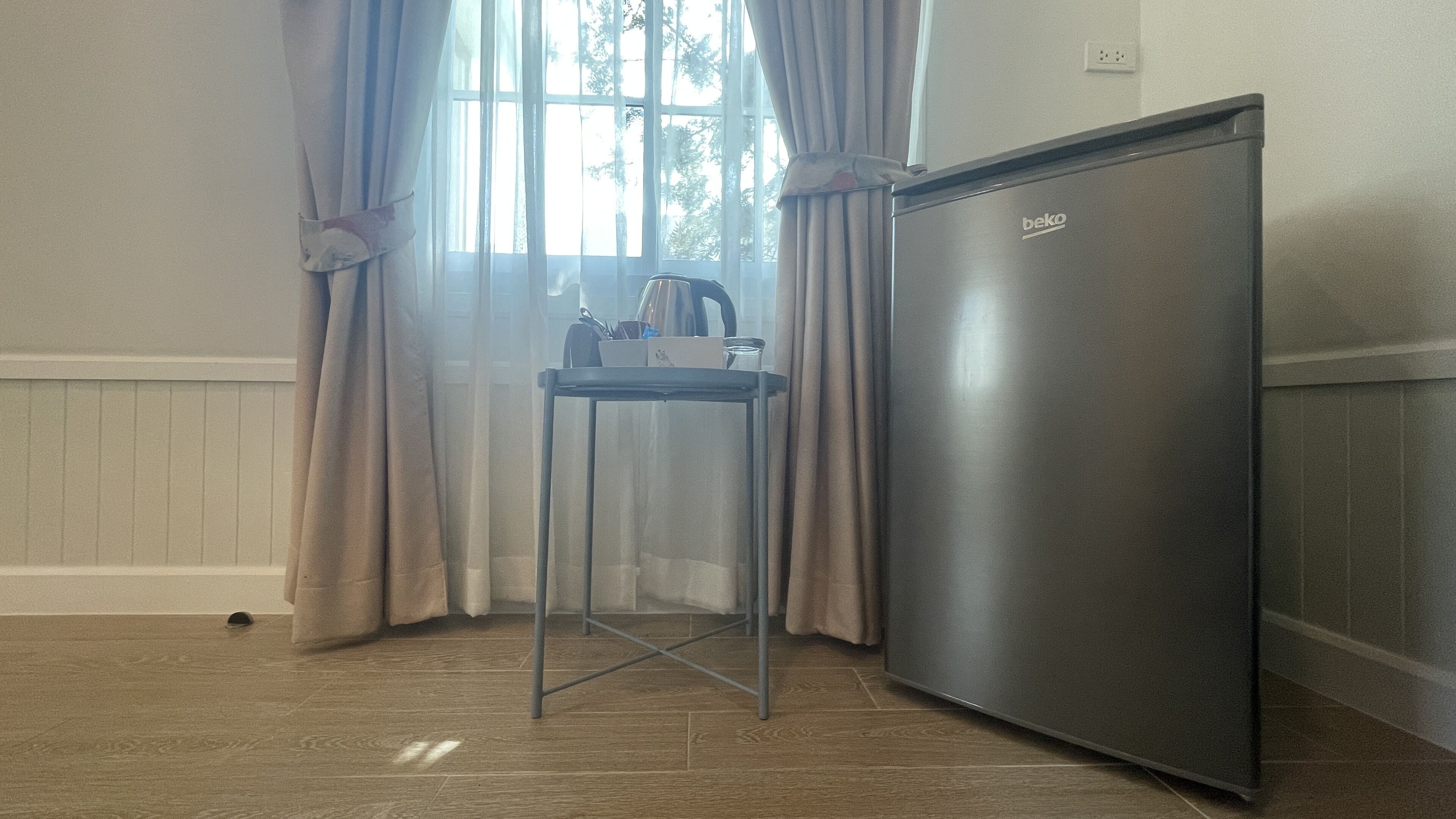 Une petite cuisine équipée d’un réfrigérateur en acier inoxydable, d’une petite table avec une bouilloire et d’une fenêtre avec des rideaux.