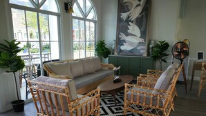 A spacious living room with a large sofa, two wicker chairs, a coffee table, and a black and white patterned rug. There are potted plants and a wall-mounted fan. - Vermont Khao Yai (Pak Chong)