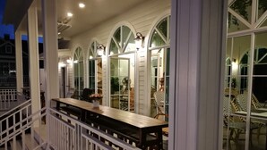 A covered outdoor dining area with a wooden table and chairs, white railings, and large arched windows. - Vermont Khao Yai (Pak Chong)