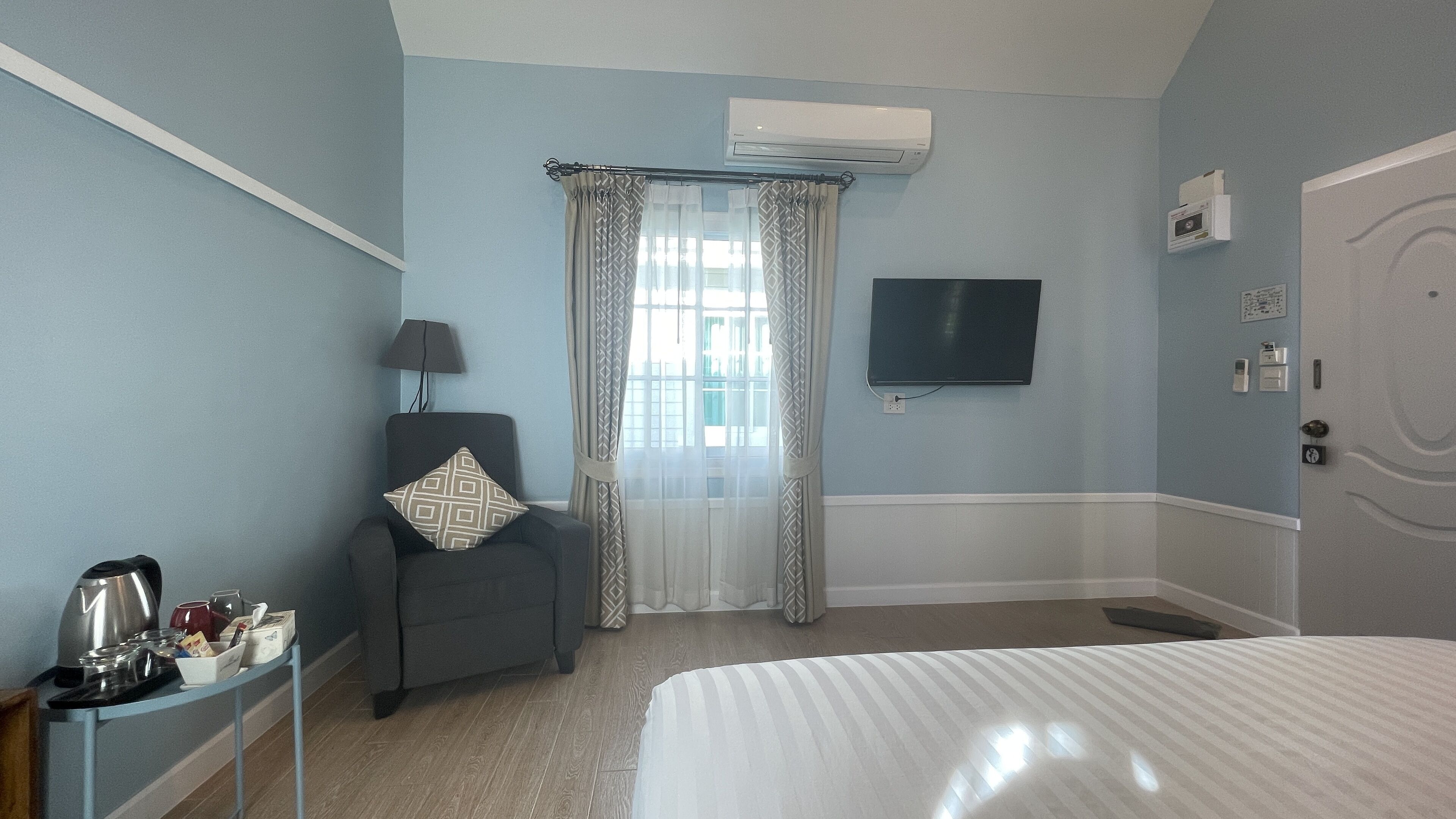 A hotel room with a bed, a chair with a patterned pillow, a small table with a kettle and cups, a TV mounted on the wall, and an air conditioning unit.
